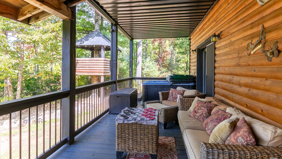 A comfortable outdoor seating area and hot tub are set on a spacious deck, framed by lush greenery. A wooden play structure for children can be seen nearby, creating a welcoming atmosphere for both relaxation and playful gatherings. The space feels cozy, communal, and closely connected to nature.