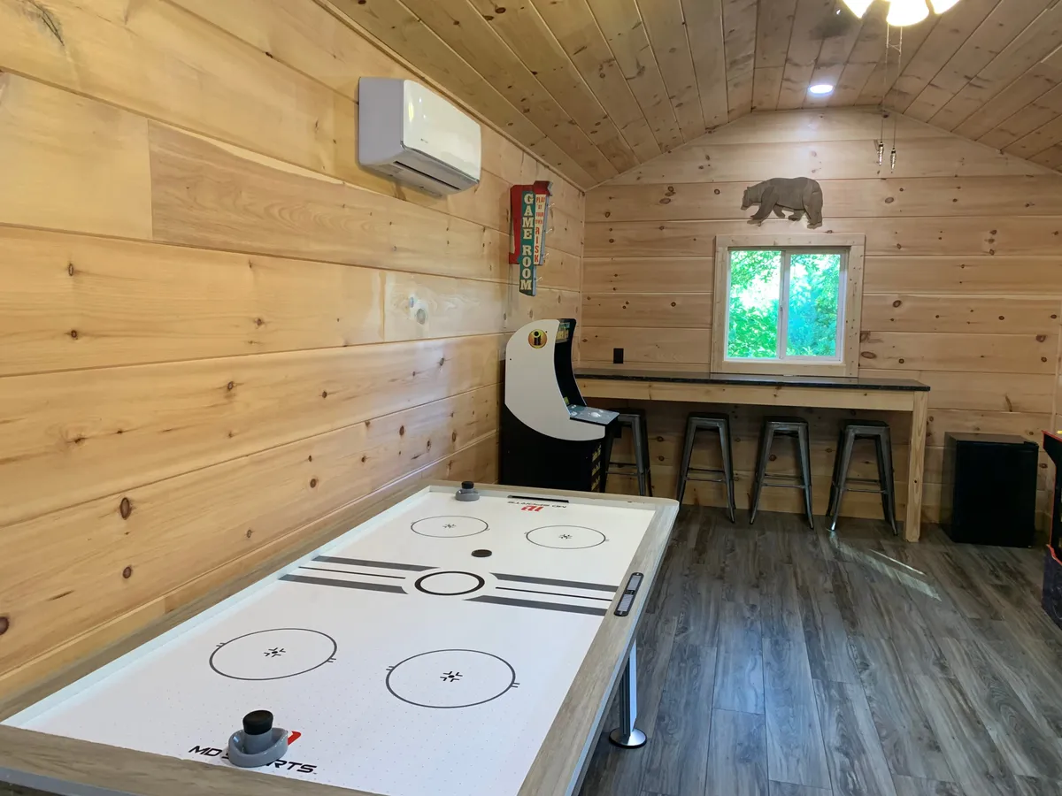 A vibrant game loft featuring an air hockey table, a classic arcade game machine, a wooden bar table with stools, and a large window looking out onto greenery. The scene is lively with ample space for socializing, playing games, and enjoying snacks—all under natural light that fills the room.