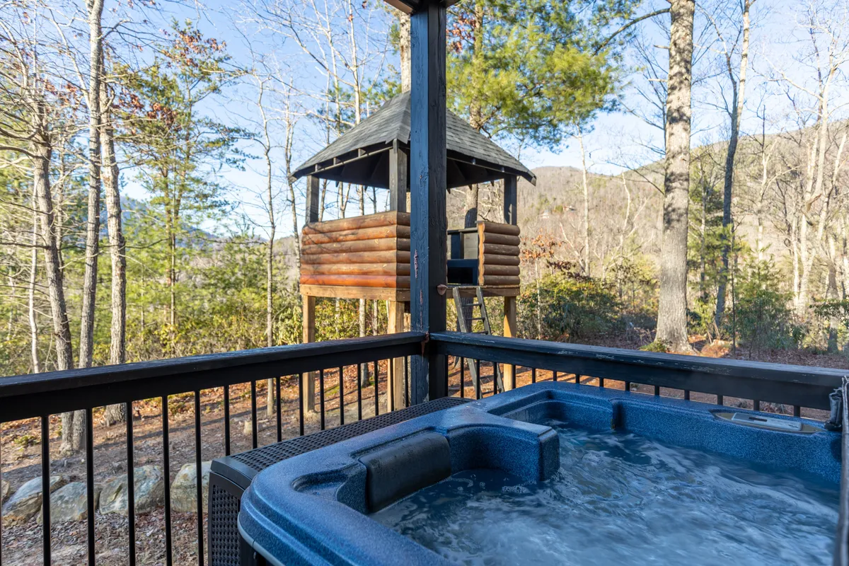 A bubbling hot tub sits on the deck, surrounded by trees, with a quaint playhouse nearby. The area is peaceful and private, inviting guests to relax in the hot tub while taking in the tranquil natural setting. The scene hints at both adult relaxation and family-friendly fun.