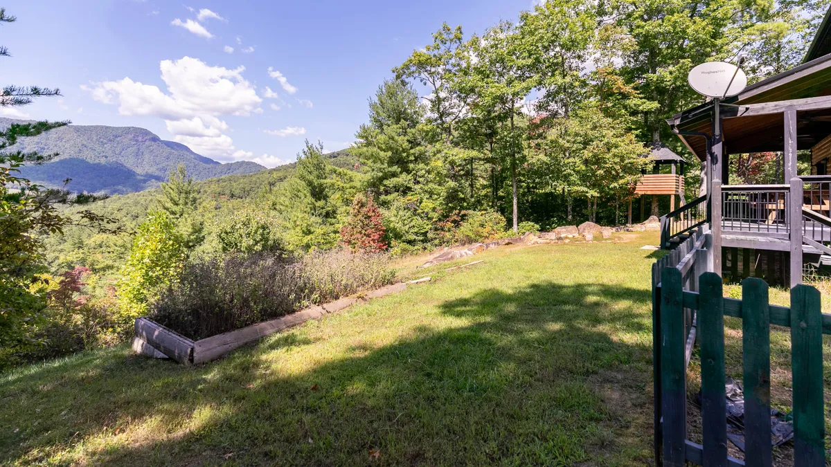 A sweeping view from the deck captures layered mountains in the distance, framed by lush greenery and a wooden fence. The deck area provides a perfect vantage point for sunrise coffee or sunset relaxation amidst the mountain magic.