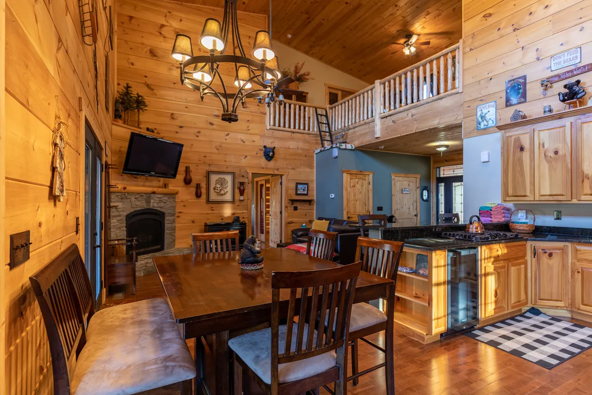 The dining area is highlighted here, featuring a large wooden dining table set beneath a chandelier. The adjacent fireplace and surrounding cabinetry reinforce the mountain lodge atmosphere. The space clearly invites group meals, board games, and lively conversations.