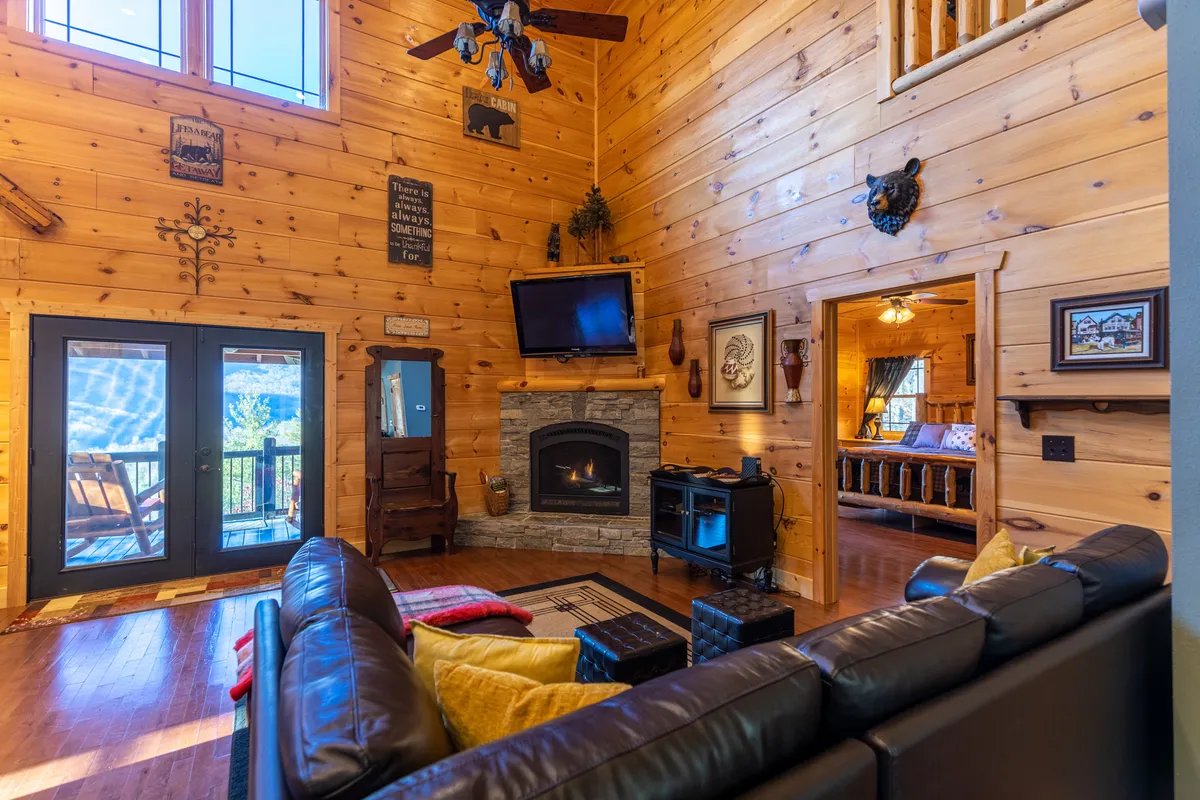 A warm living room centers around a stunning stone fireplace, flanked by a leather sofa, large windows, and a wooden ceiling. The space is bathed in natural light, with decorative wall art adding to the inviting mountain ambiance. The arrangement encourages curling up by the fire for intimate conversations or relaxation.