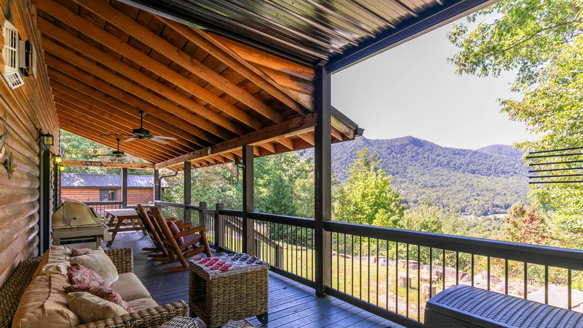 The image showcases the inviting exterior porch of a cozy log cabin with classic wooden rocking chairs and a woven sofa positioned to overlook breathtaking mountain vistas. Sturdy wooden beams frame the spacious porch, creating a serene spot ideal for relaxing, reading, or gathering with friends. The peaceful mountain setting hints at quiet mornings watching the sunrise and memorable evenings spent stargazing.