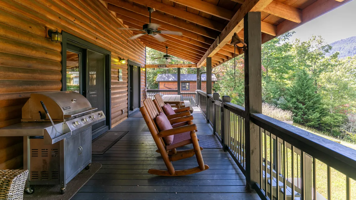 A cozy exterior space featuring classic rocking chairs and a grill beneath sturdy wooden beams. The view extends into the forest, offering a peaceful retreat for relaxation, socializing, and outdoor meals as the sun sets.