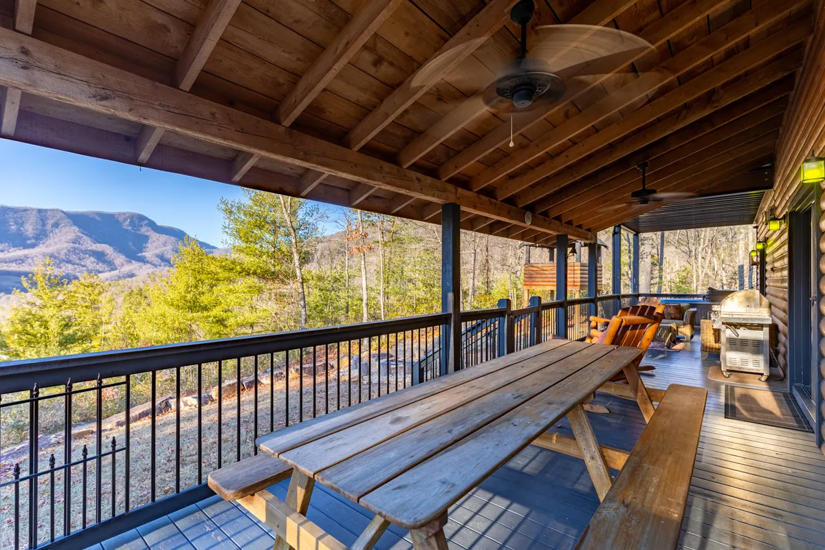 A large wooden table is set on a covered deck, surrounded by comfortable seating and a grill. The wooden ceiling overhead, along with the open-air design, frames sweeping mountain views in the distance. The space feels communal and inviting, ideal for group gatherings or quiet meals with nature as the backdrop.