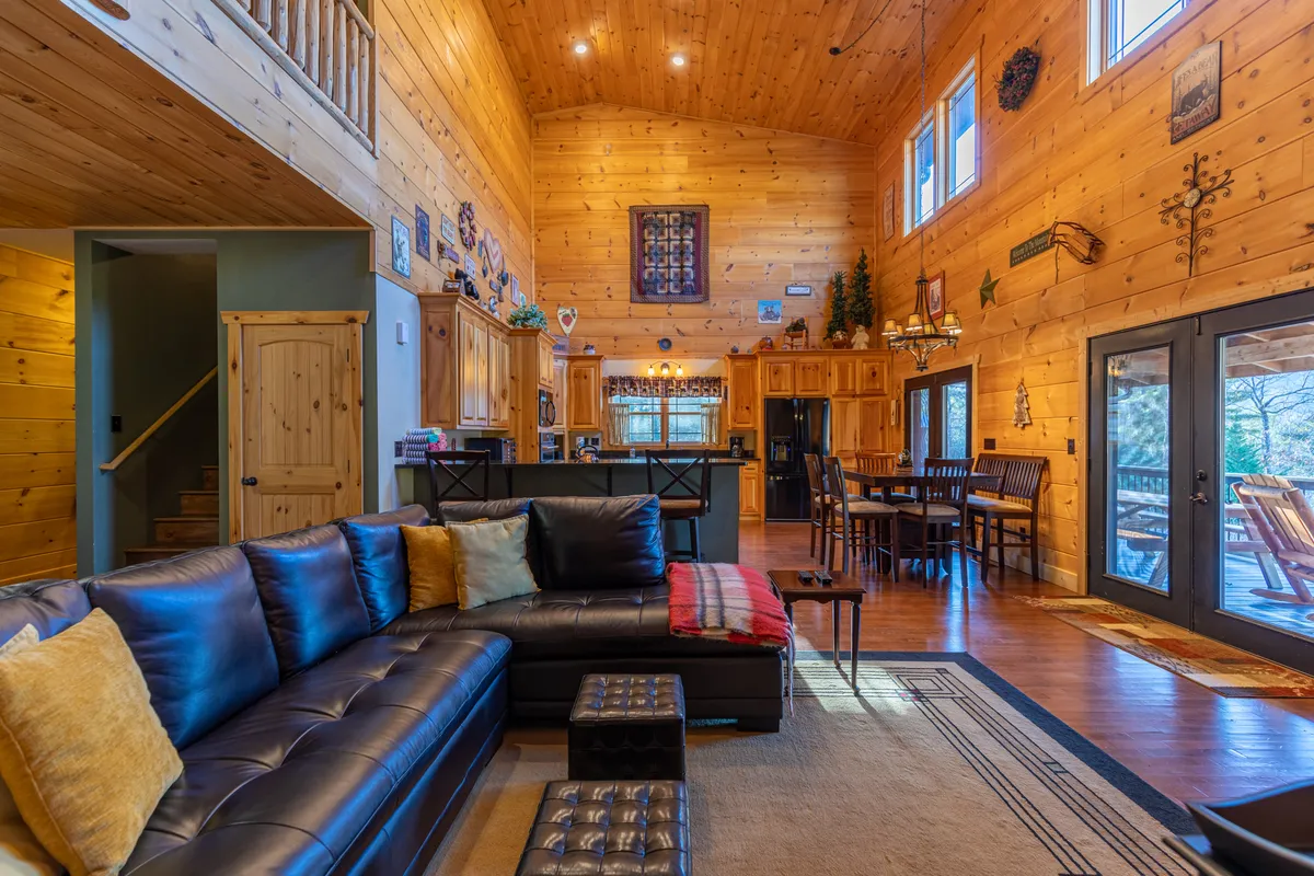 This image features an expansive open-concept living area with a large sectional sofa perfectly positioned for family gatherings. The scene also highlights warm wooden kitchen cabinetry and a visible dining area, with large windows that let in abundant natural light and offer tranquil views of the surrounding nature. The overall mood is cozy and inviting, capturing the essence of a communal mountain retreat.