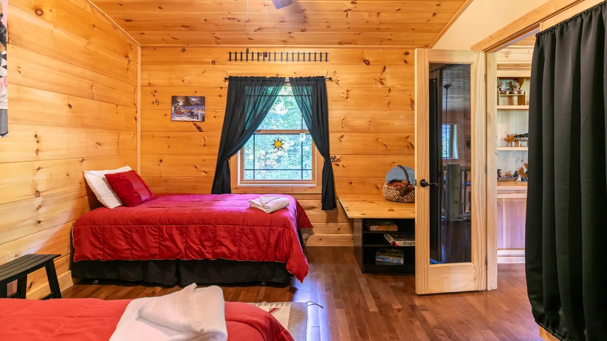 Featuring two beds with vibrant red bedding, wood-paneled walls, a large window with curtains, and a small desk, this room feels both comfortable and home-like. The space is bathed in soft natural light, encouraging relaxation and togetherness.